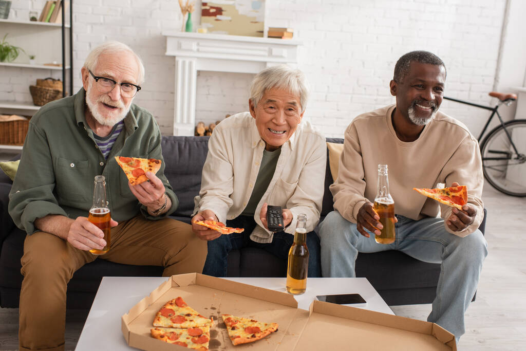 positivo interracial sênior amigos segurando pizza e cerveja enquanto assistindo tv em casa  - Foto, Imagem