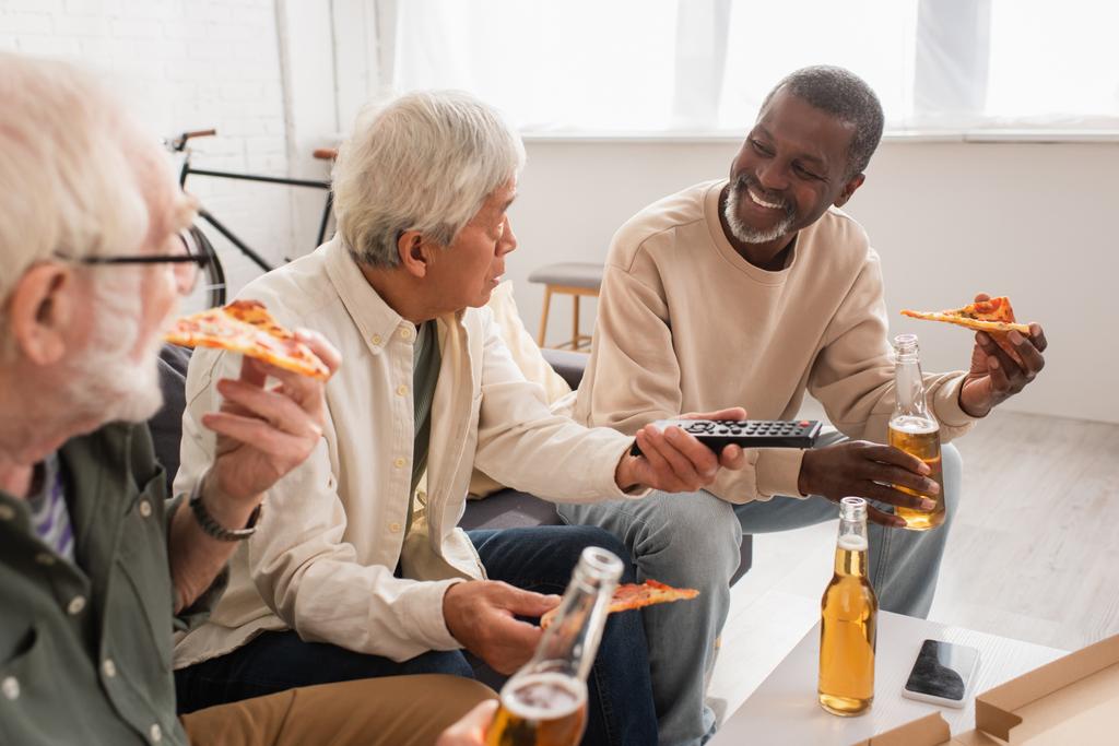 アフリカ系アメリカ人の男がビールとピザを持っています多民族の友人の近くにリモコンで自宅で  - 写真・画像