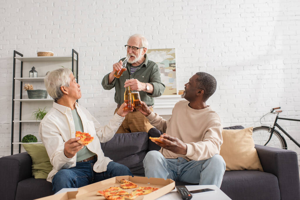 Ancianos amigos multiétnicos tintineo cerveza cerca de pizza y mando a distancia en casa  - Foto, Imagen