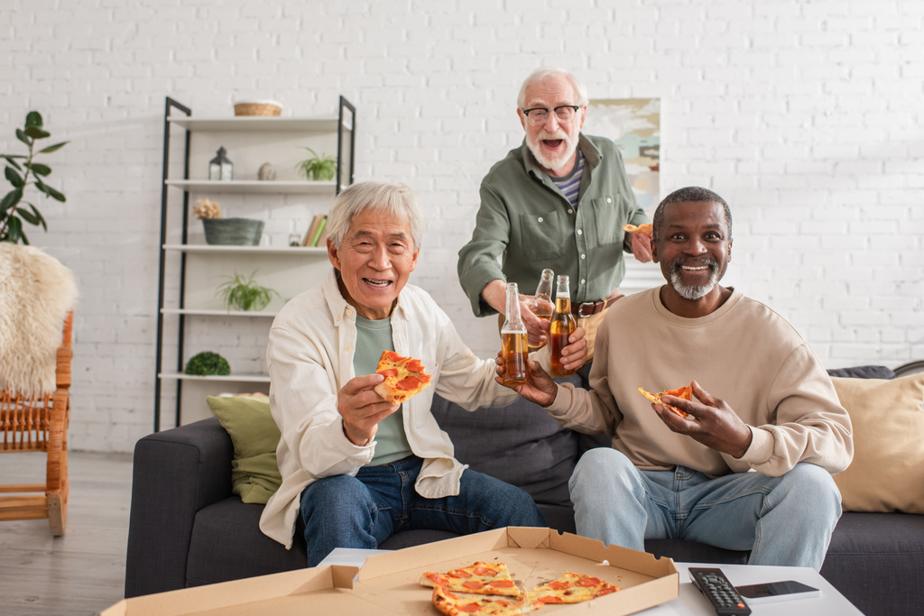 Heureux aînés multiethniques tenant des bouteilles de pizza et de bière à la maison  - Photo, image