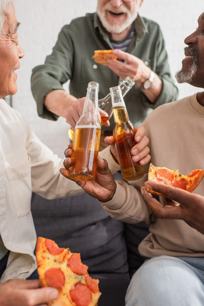 Amigos mayores interraciales sosteniendo botellas de cerveza y pizza en casa  - Foto, Imagen
