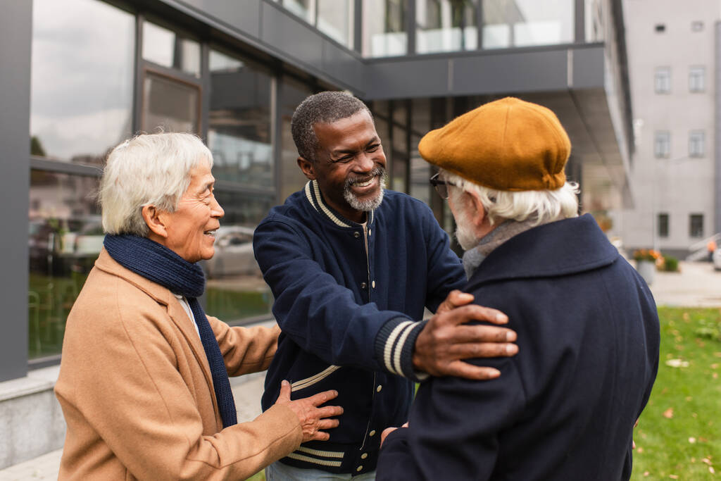 feliz africano americano pensionista abrazando senior amigo cerca asiático hombre en urbano calle  - Foto, imagen