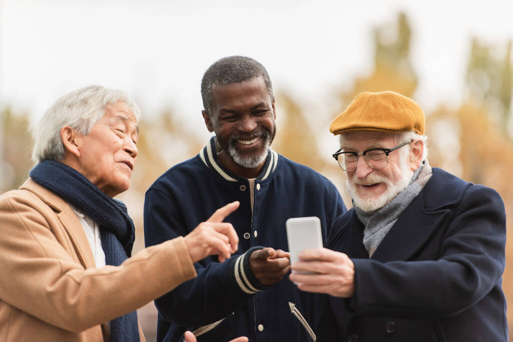 Pozytywni emeryci międzyrasowi wskazujący na smartfona na ulicy miejskiej  - Zdjęcie, obraz
