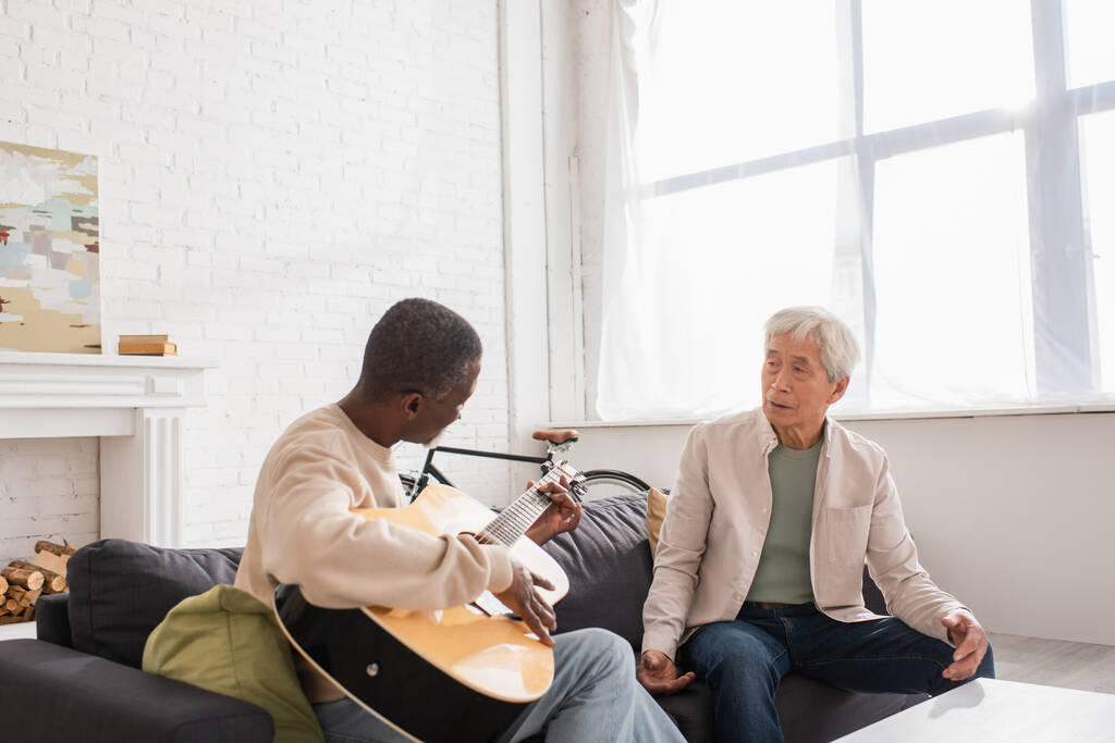 異人種間の年金受給者が自宅でソファでアコースティックギターを演奏  - 写真・画像