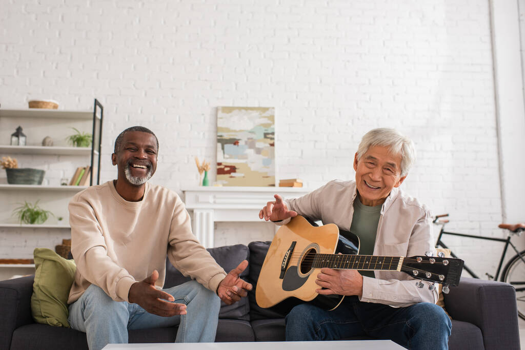 Веселые мультиэтнические пенсионеры с акустической гитарой, смотрящие в камеру на диване дома  - Фото, изображение
