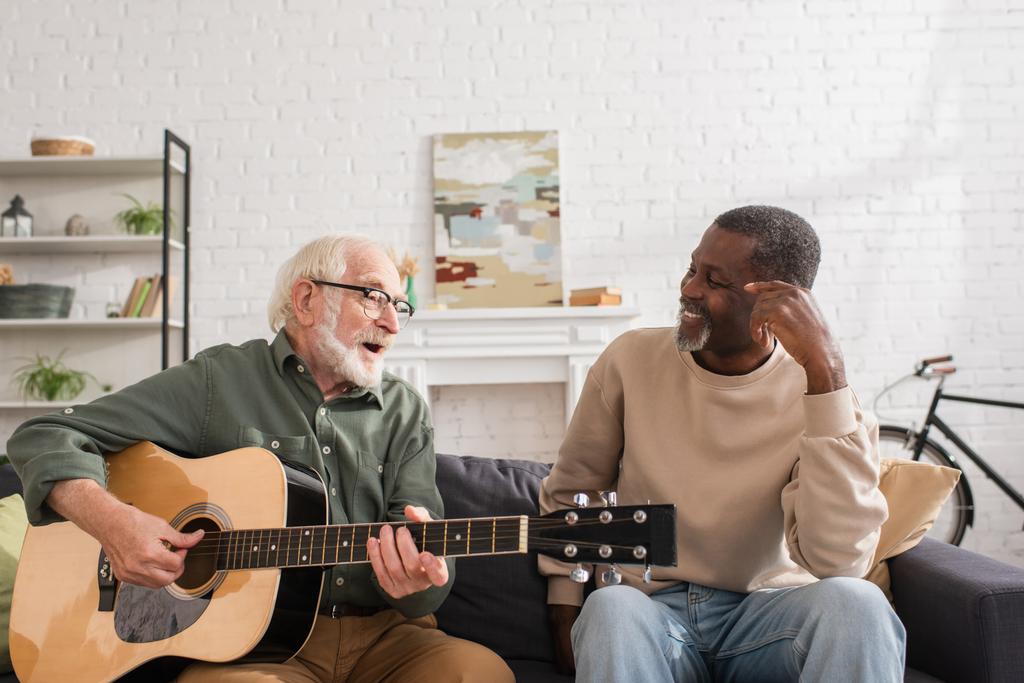 Hombre mayor tocando la guitarra acústica cerca de sonreír amigo afroamericano en el sofá en casa  - Foto, imagen