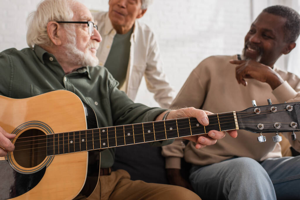 Guitarra acústica en manos de un hombre mayor tocando cerca de amigos multiétnicos en casa  - Foto, Imagen