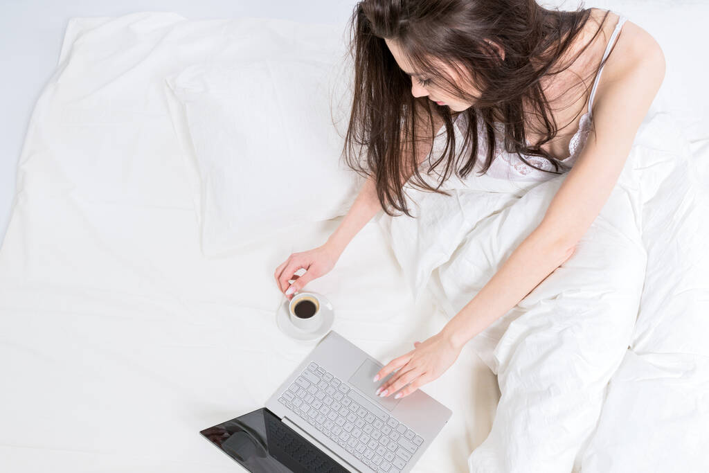 ノートパソコンとコーヒーカップの横にある白いベッドの上に可愛いブルネットが座っています。おはようございます。家からの仕事. - 写真・画像