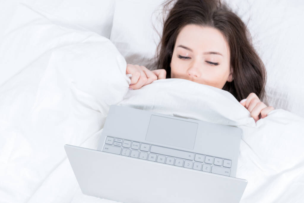 A morena tem medo de olhar para o computador portátil. A menina jaz na cama, coberta com um cobertor, um laptop está em cima dela. - Foto, Imagem
