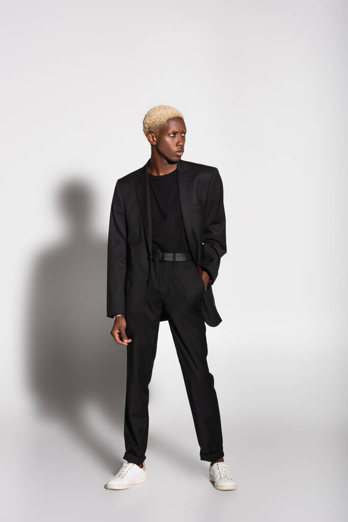 vista integrale dell'uomo afroamericano in completo nero e sneakers bianche in piedi con mano in tasca su grigio con ombra - Foto, immagini