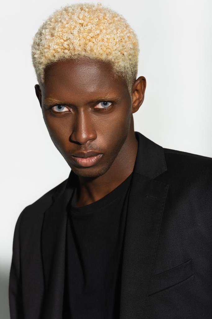 junger afrikanisch-amerikanischer Mann mit blonden Haaren blickt isoliert in die Kamera auf grau - Foto, Bild