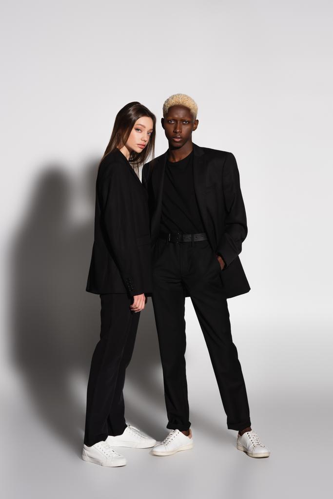 Ansicht der blonden afrikanisch-amerikanischen Mann und brünette Frau in schwarzen Anzügen auf grau mit Schatten - Foto, Bild
