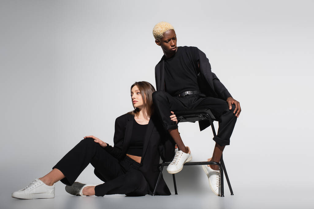 stylish woman on floor and blonde african american woman on chair posing in black suits on grey with shadow - Photo, Image