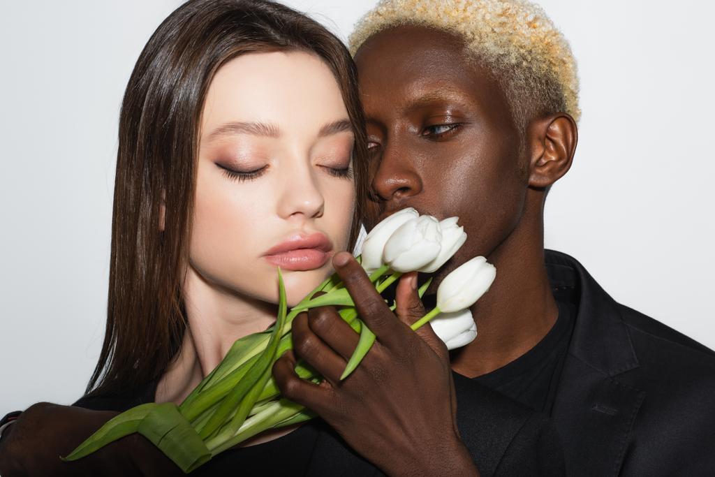 blonde afrikanisch-amerikanische Mann hält weiße Tulpen neben hübsche Frau mit geschlossenen Augen isoliert auf grau - Foto, Bild