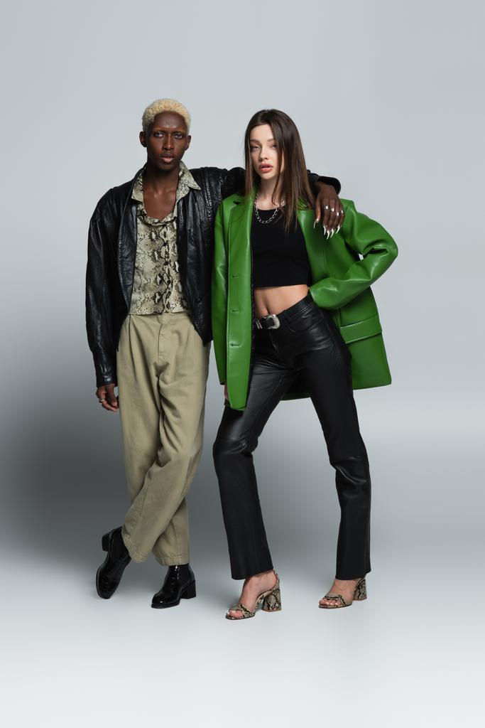 visão de comprimento total de casal interracial elegante em jaquetas de couro olhando para a câmera no cinza - Foto, Imagem