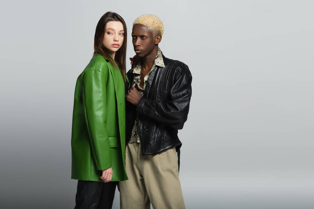 bionda afroamericana uomo e bruna donna in giacche di pelle in piedi sul grigio - Foto, immagini