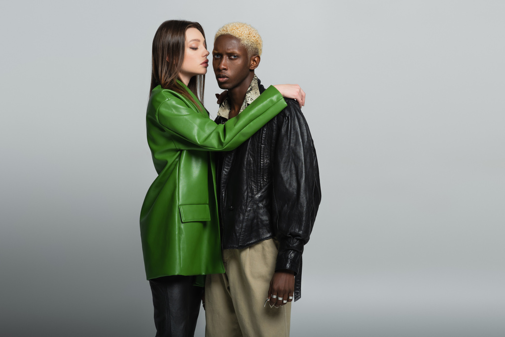 брюнетка в зеленій шкіряній куртці обіймає блондинку афроамериканця на сірому
 - Фото, зображення