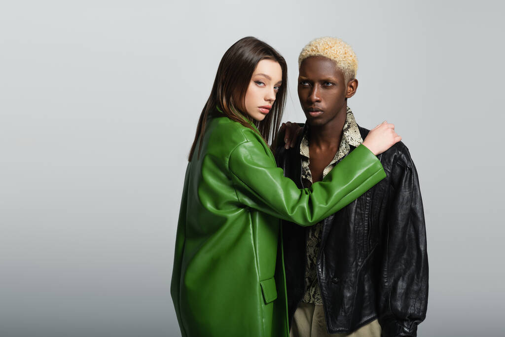 чувственная женщина в зеленой кожаной куртке, смотрящая в камеру и обнимая блондинку африканского мужчины, изолированного на сером - Фото, изображение