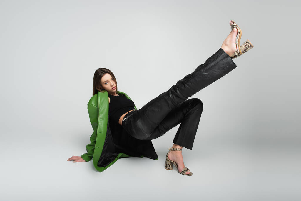 stylische Frau in Lederkleidung und Sandalen sitzend mit erhobenem Bein auf grau - Foto, Bild