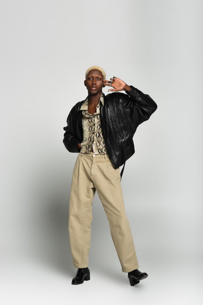 visão de comprimento total de homem americano africano loiro em jaqueta preta e calças bege olhando para a câmera em cinza - Foto, Imagem