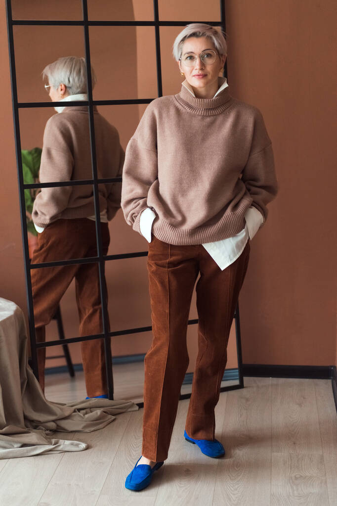 mujer de moda en suéter marrón de gran tamaño y pantalones de pie en el lugar de trabajo moderno u oficina - Foto, imagen