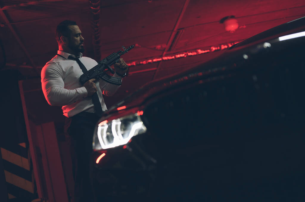 pistoolwet concept van sterke jonge bebaarde gangster man in wit shirt en stropdas holding machine pistool in de buurt van sport auto in rood verlichte undeground garage - Foto, afbeelding