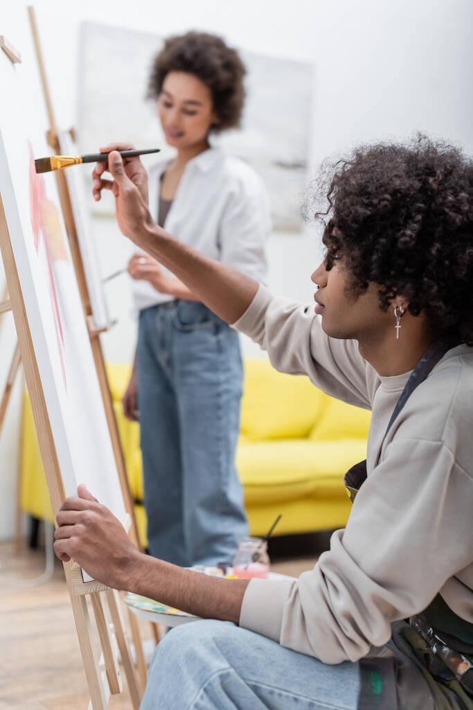 Jovem afro-americano em avental pintura sobre tela perto de namorada borrada em casa  - Foto, Imagem