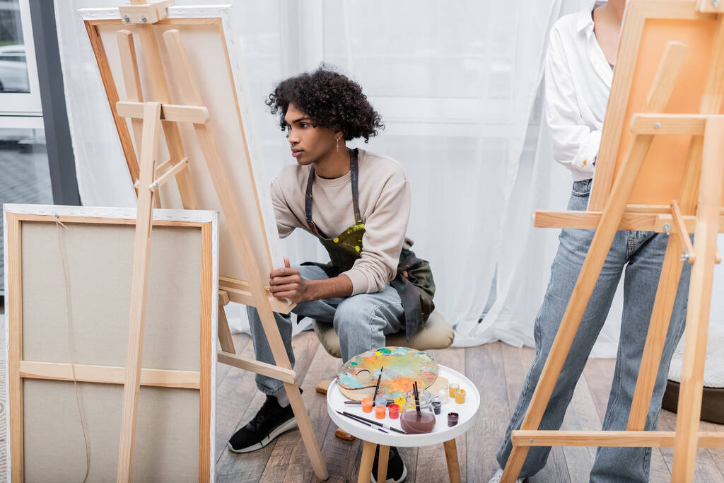Молодий афроамериканець у фартусі малює на полотні поруч з фарбами та дівчиною вдома.  - Фото, зображення