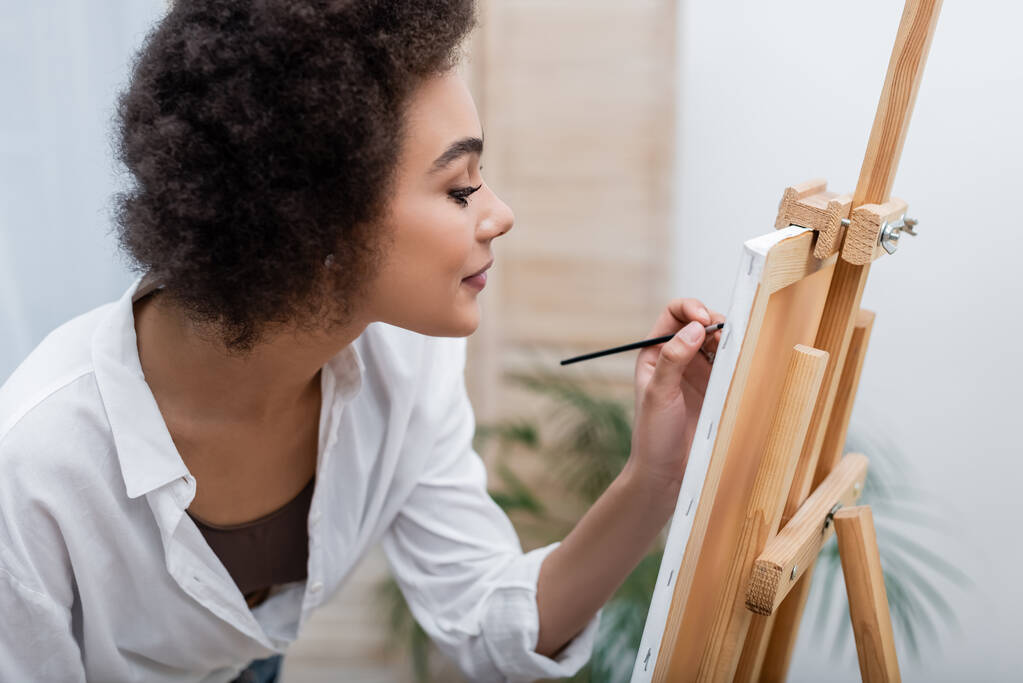 Vista lateral de la mujer afroamericana pintando sobre lienzo en sala de estar   - Foto, Imagen