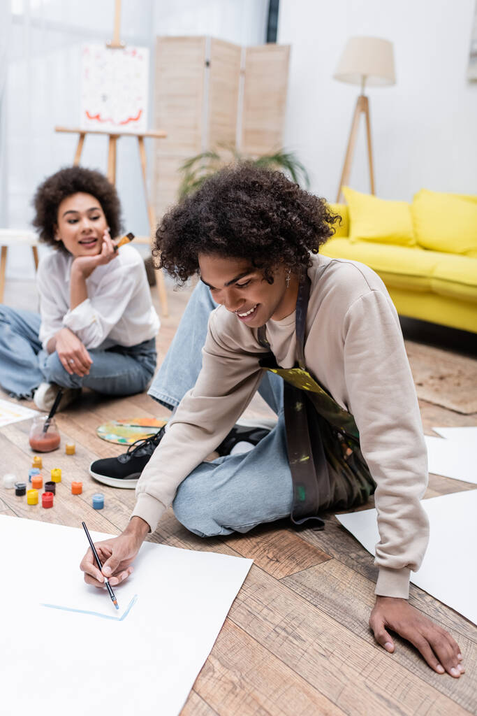 Smiling african american man painting on floor near blurred girlfriend at home  - Photo, Image