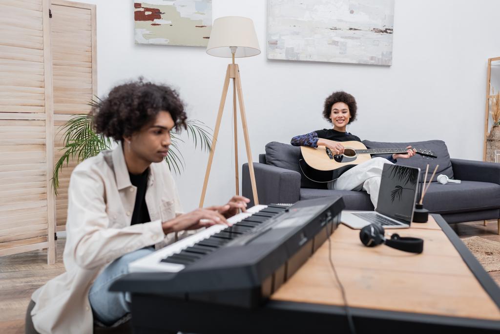 African american woman playing acoustic guitar near blurred boyfriend with synthesizer and laptop at home  - Photo, Image