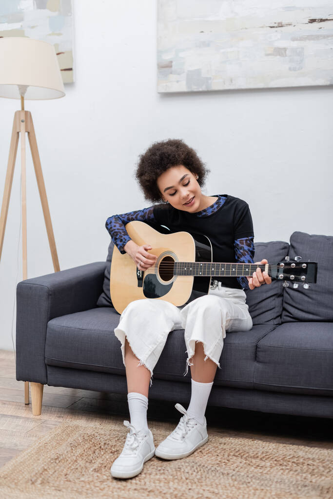 Mosolygó afro-amerikai nő játszik akusztikus gitár kanapén otthon  - Fotó, kép
