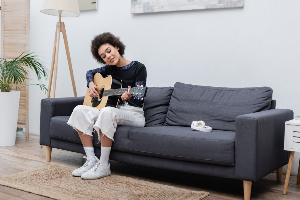 Mujer afroamericana joven tocando la guitarra acústica cerca de los auriculares en el sofá  - Foto, imagen