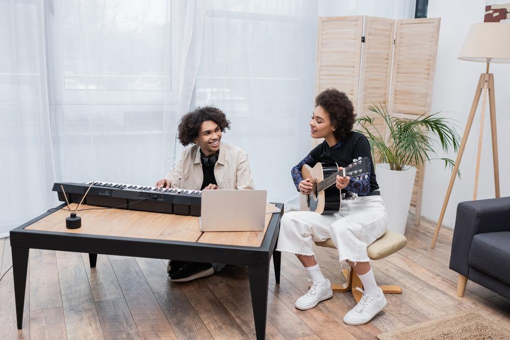Smiling african american woman playing acoustic guitar near boyfriend with synthesizer and laptop in living room  - Photo, Image
