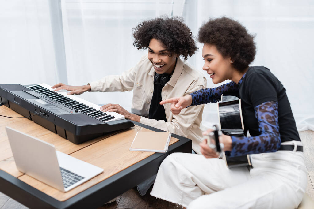 Glimlachende Afrikaanse Amerikaanse vrouw met akoestische gitaar wijzen met de vinger in de buurt vriendje spelen synthesizer en laptop thuis  - Foto, afbeelding