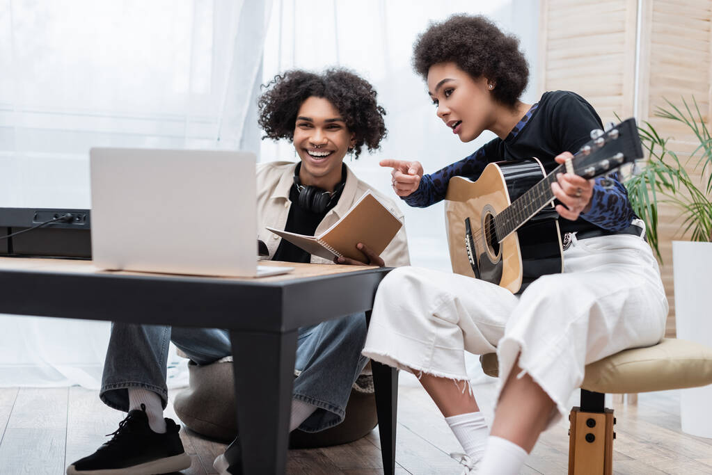 Sonriente hombre afroamericano mirando borrosa portátil cerca sintetizador y novia con guitarra acústica en casa  - Foto, imagen