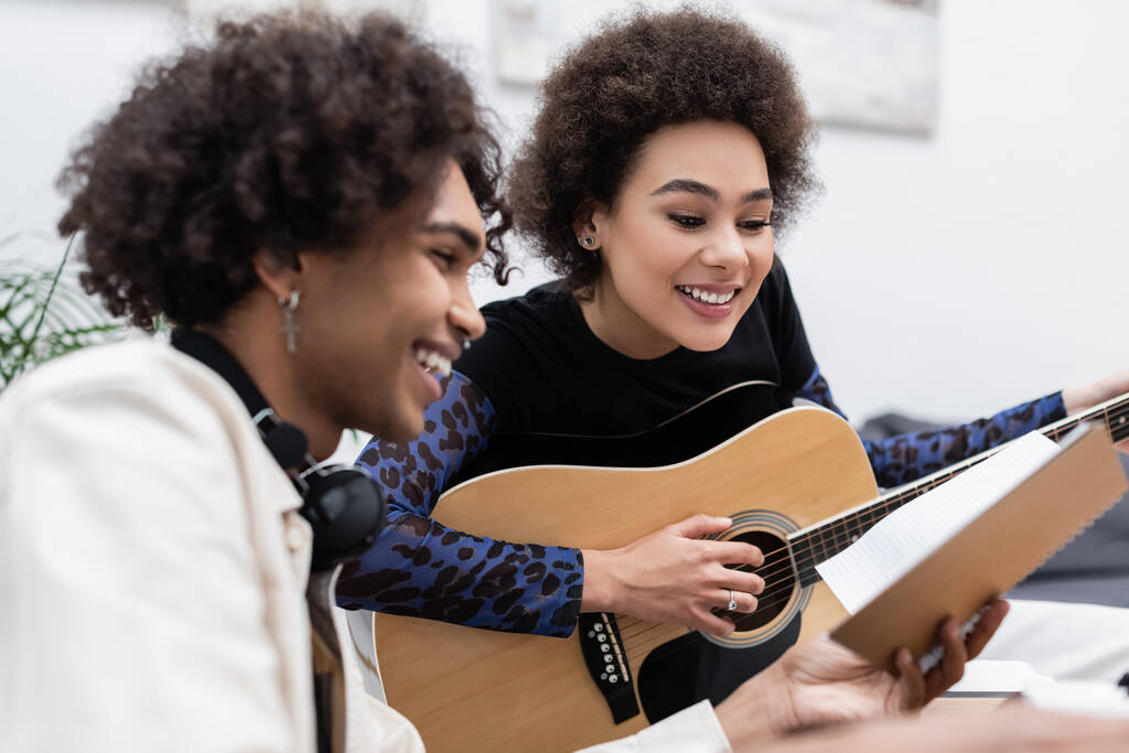 Jovem afro-americano segurando notebook perto namorada tocando guitarra acústica em casa  - Foto, Imagem