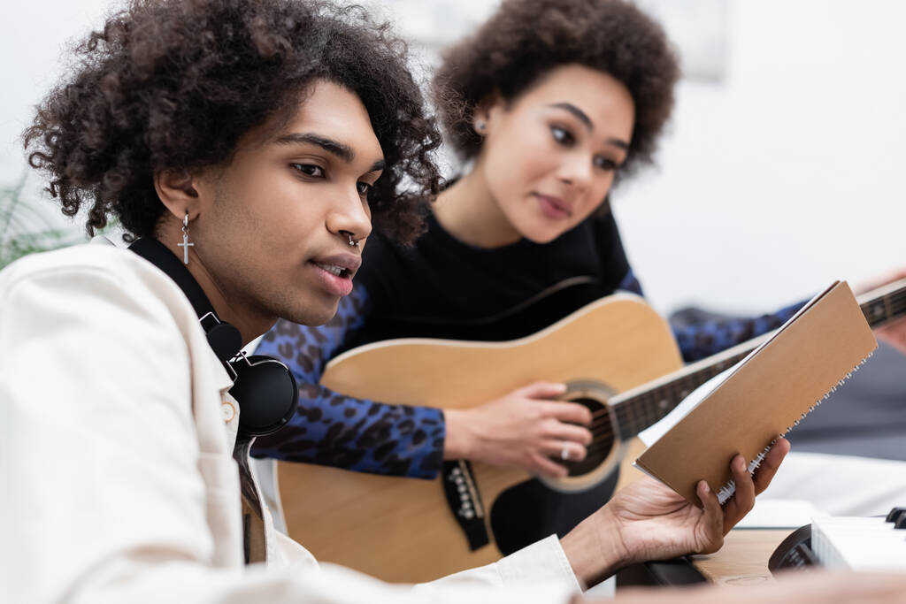 Africano americano com fones de ouvido segurando notebook perto namorada borrada tocando guitarra acústica e sintetizador em casa  - Foto, Imagem