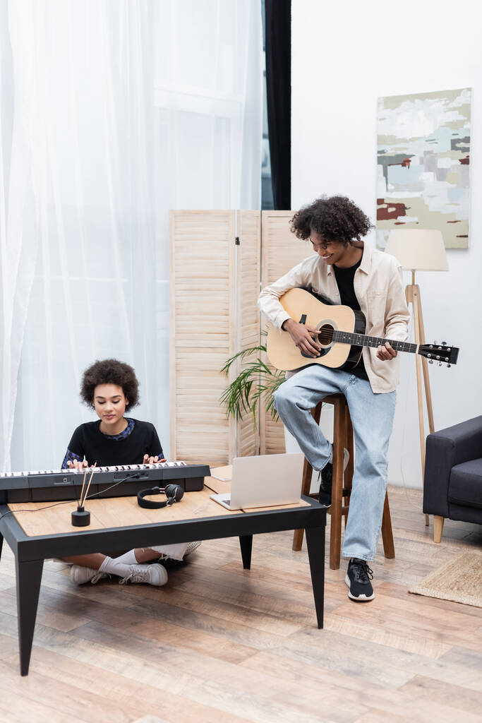 Souriant homme afro-américain jouant de la guitare acoustique près de petite amie avec synthétiseur et ordinateur portable à la maison  - Photo, image