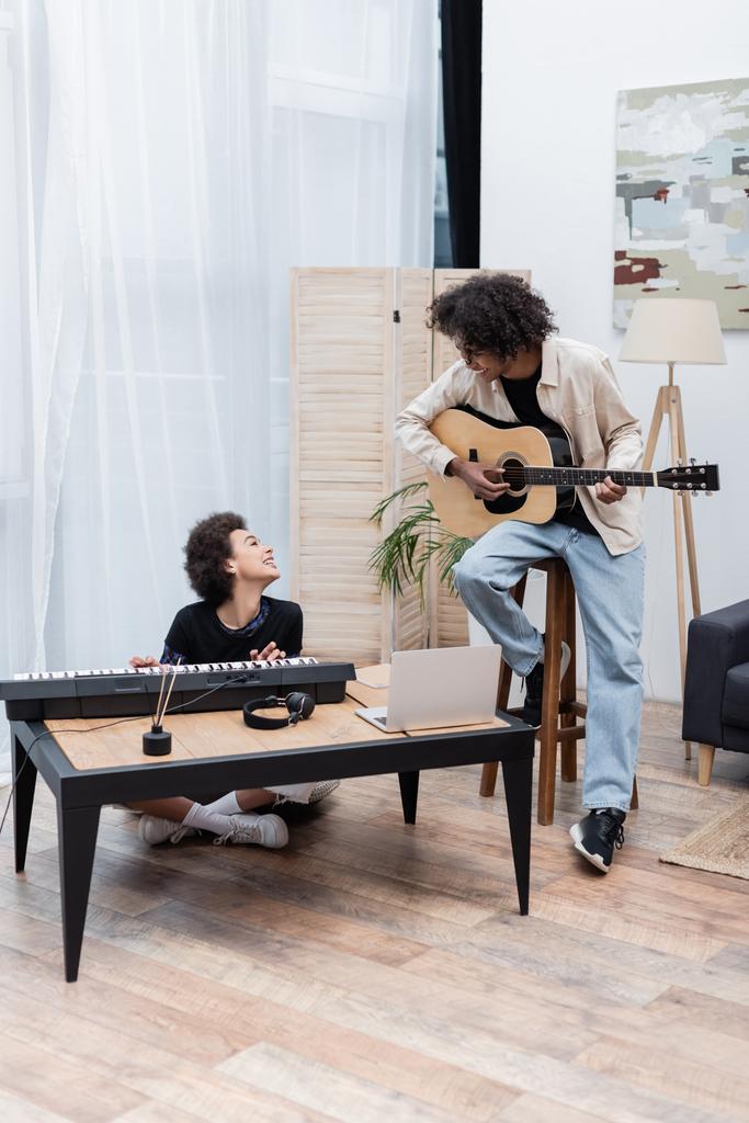 Χαμογελώντας αφροαμερικάνικο ζευγάρι κοιτάζοντας ο ένας τον άλλο ενώ παίζουν μουσικά όργανα κοντά στο φορητό υπολογιστή στο σπίτι  - Φωτογραφία, εικόνα