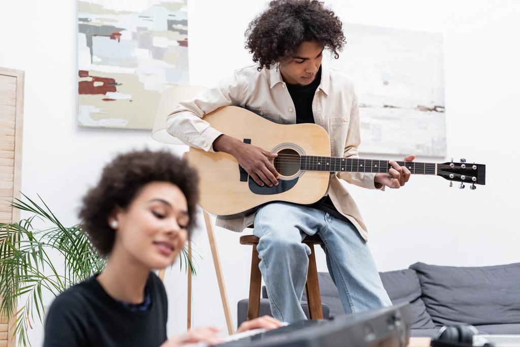 Hombre afroamericano tocando la guitarra acústica cerca de novia borrosa con sintetizador en casa  - Foto, imagen