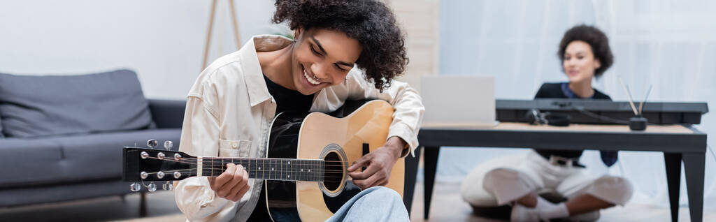 Mladý africký Američan hrající na akustickou kytaru poblíž rozmazané přítelkyně v obývacím pokoji, banner  - Fotografie, Obrázek