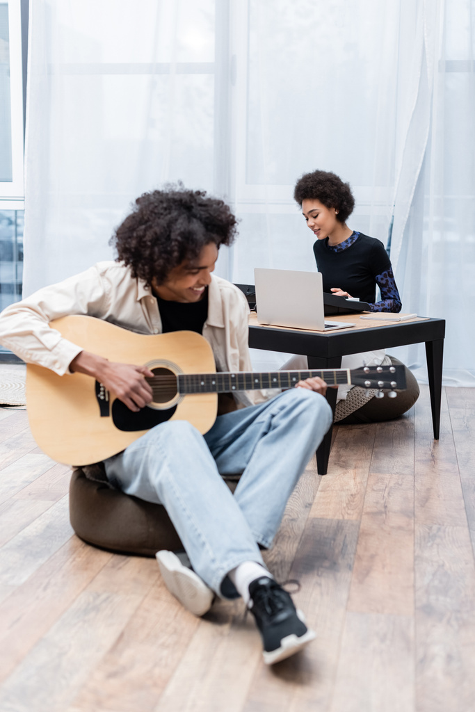 Gülümseyen Afro-Amerikan kadın dizüstü bilgisayarın yanında sentezleyici oynuyor ve erkek arkadaşı oturma odasında akustik gitarlı.  - Fotoğraf, Görsel