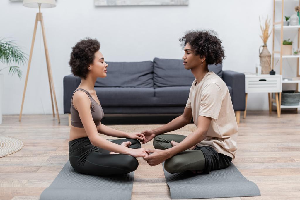 Afro-Amerikalı genç çiftin el ele tutuşup evdeki paspaslar üzerinde meditasyon yapmasının yan görüntüsü.  - Fotoğraf, Görsel