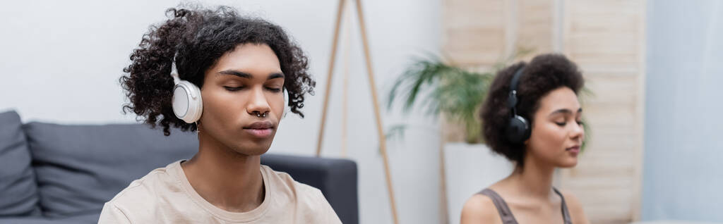 Jovem afro-americano em fones de ouvido meditando perto de namorada borrada em casa, banner  - Foto, Imagem