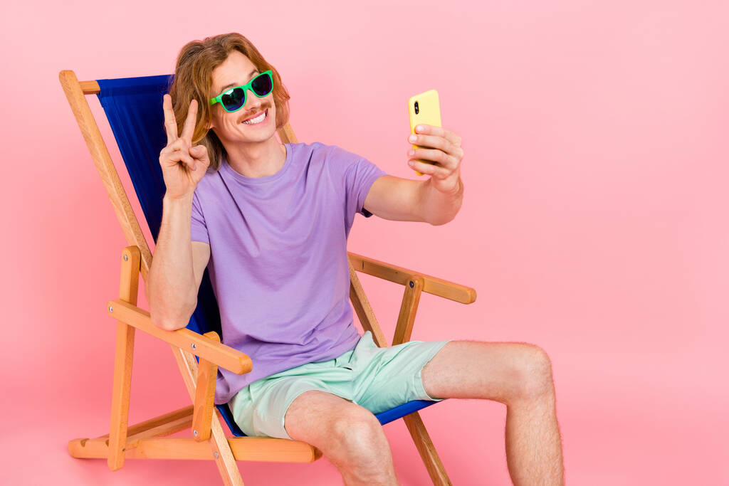 Yakışıklı, modaya uygun neşeli adamın portresi sandalyede oturup selfie çekerken pembe pastel arka planda izole edilmiş v-işareti gösteriyor. - Fotoğraf, Görsel