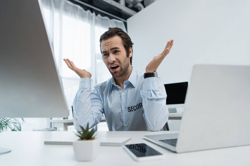 displeased security man gesturing near computer monitor and laptop in office - Photo, Image