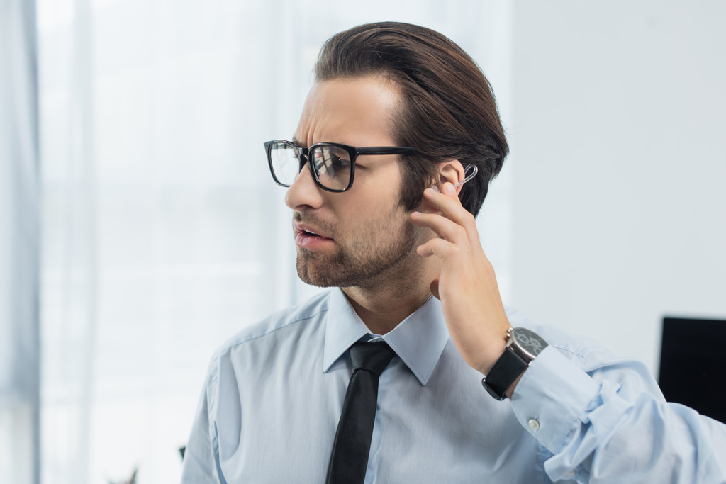 seriózní bezpečnostní muž v brýlích dotýkání sluchátka při práci v kanceláři - Fotografie, Obrázek
