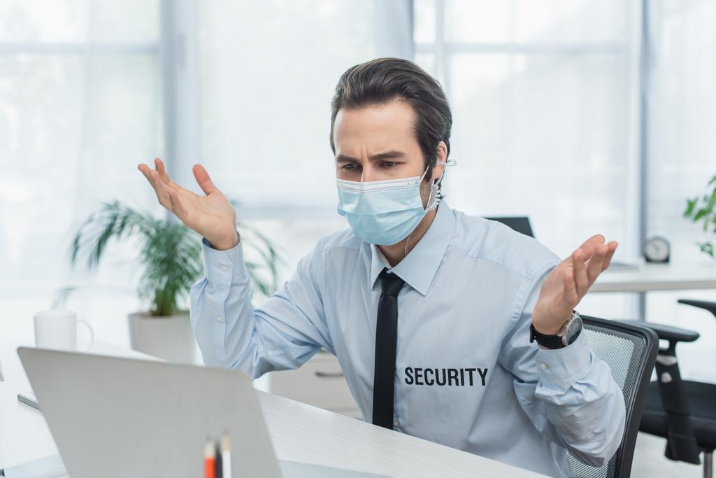 Entmutigter Security-Mann mit medizinischer Maske gestikuliert in der Nähe eines verschwommenen Laptops - Foto, Bild