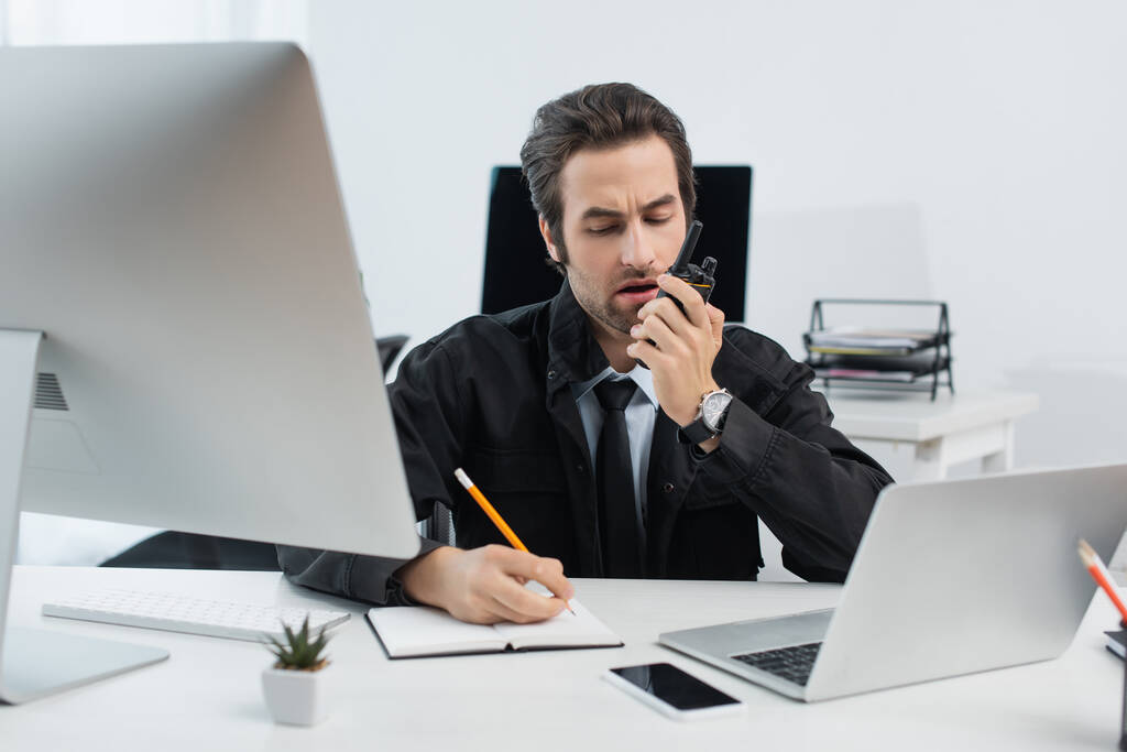 homem de segurança falando sobre walkie-talkie e escrevendo em notebook perto de computadores no escritório - Foto, Imagem